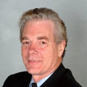 The headshot is the image of an older white man, appears to be of incongruent age, with grey-white thin hair, smiles witho...